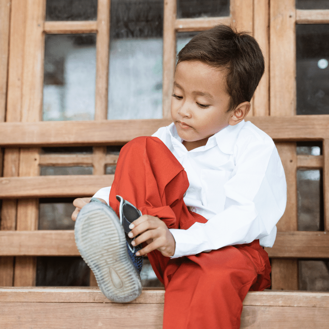 Image: A young boy in a white shirt and red pants sitting on a bench putting on his boots to illustrate the importance of teaching skills.