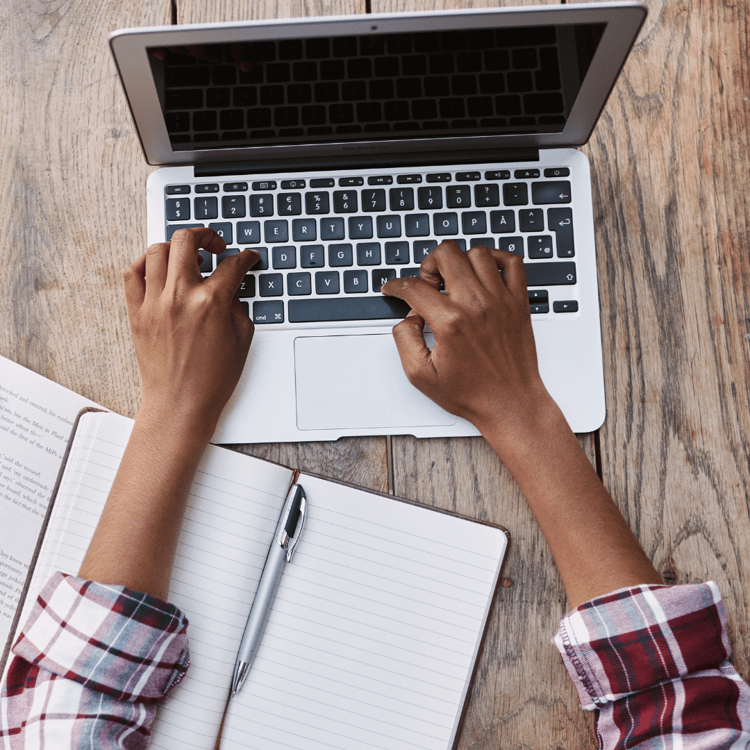 Person with rolled-up sleeves typing on a laptop keyboard, with a notebook and pen in front.