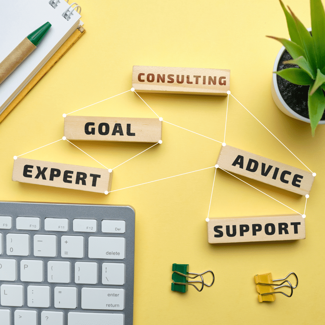 Desk with a yellow background featuring a keyboard, notebook with a pen, paper clip, and words like 'expert,' 'goal,' 'consulting,' 'advice,' and 'support.' A plant sits on one side, symbolizing the workspace of a behaviour support practitioner.