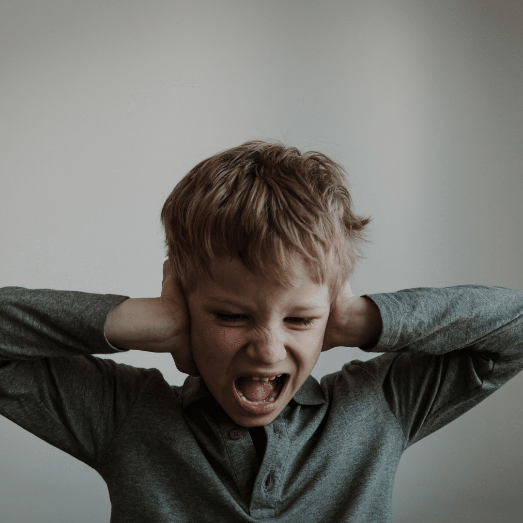 Image is of young child with hands over his ears and wide open mouth looking distressed. This illustrates how setting events can be overwhelming and contribute to behaviours of concern.